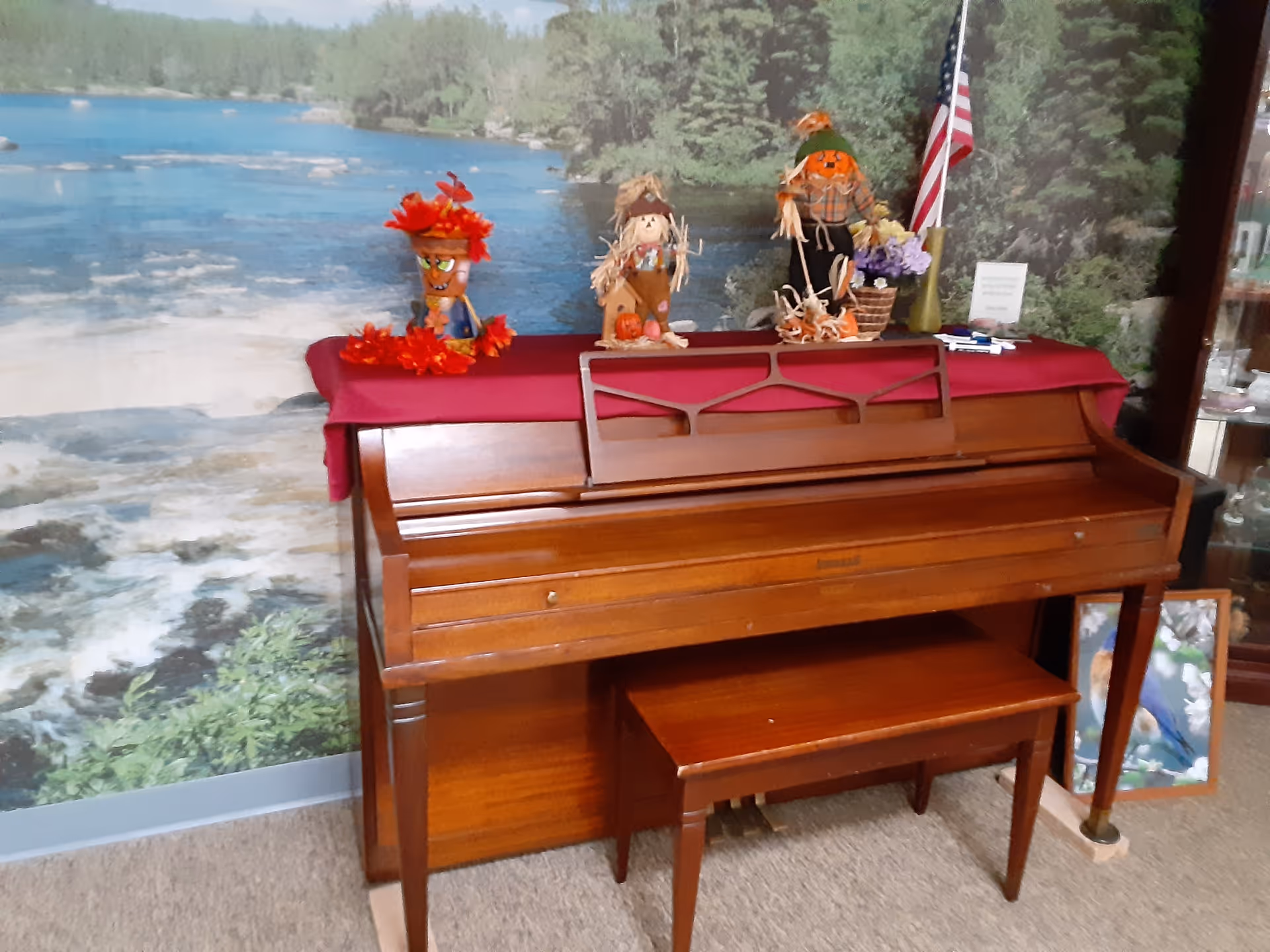 A wooden upright piano with a matching bench in front of a wall mural depicting a river and forest scene. The piano top is covered with a red cloth and decorated with three small scarecrow figurines, a basket of flowers, and an American flag. There is a framed picture leaning against the wall to the right of the piano.