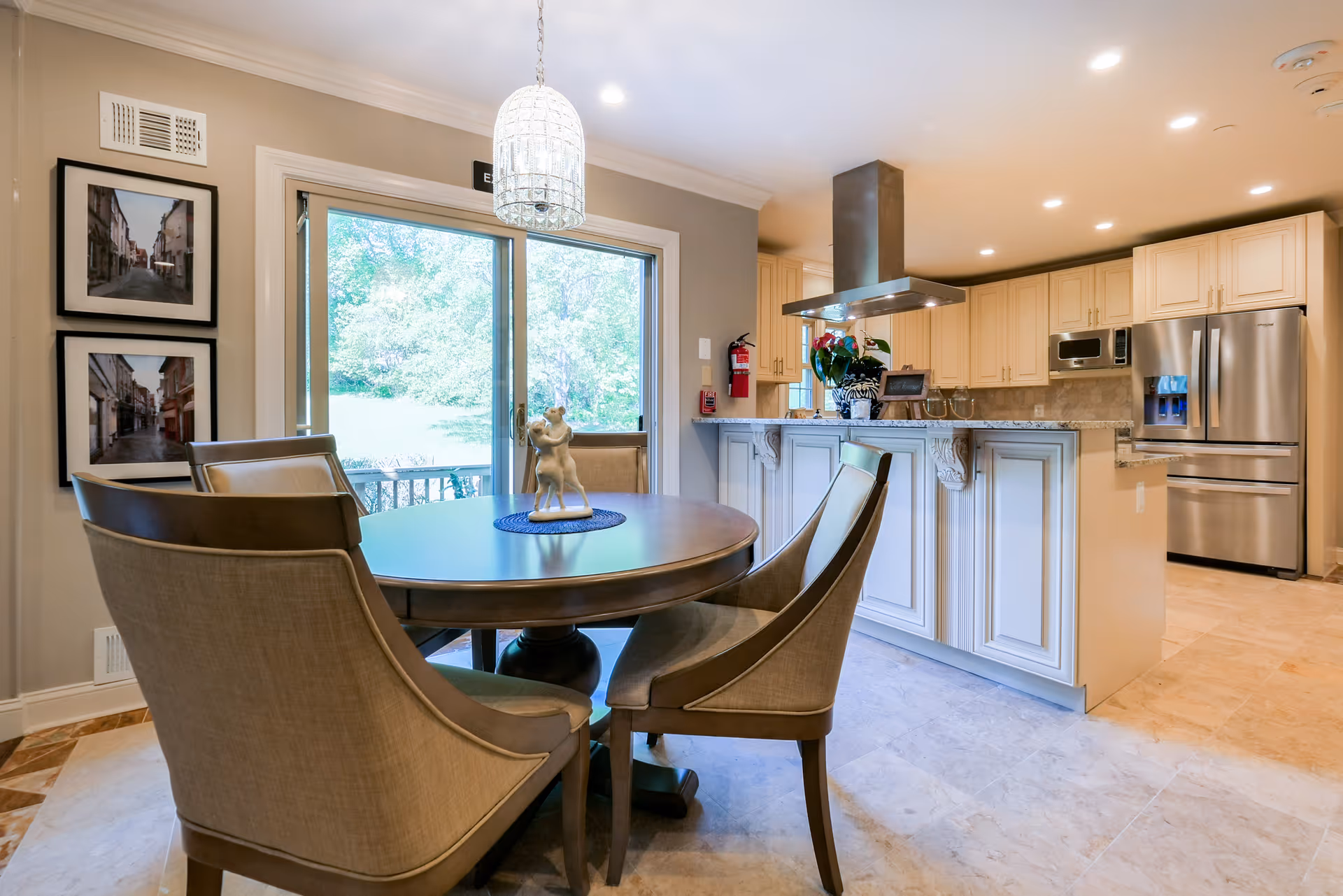 A bright and modern kitchen and dining area featuring a round wooden dining table with four upholstered chairs. A decorative sculpture is placed on the table. The kitchen has cream-colored cabinets, stainless steel appliances including a refrigerator and microwave, and a granite countertop with a built-in range hood. Large sliding glass doors provide a view of a green outdoor space. Two framed pictures hang on the wall near the door.