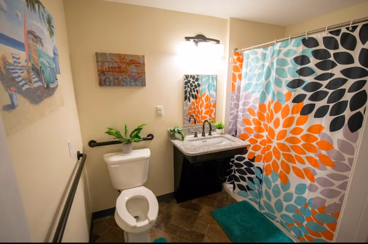 A bathroom with a white toilet, a sink with a granite countertop, a mirror above the sink, and a colorful shower curtain featuring a floral pattern in orange, black, teal, and purple. There are two green plants on the sink and toilet tank, a teal bath mat on the floor, and two wall decorations including a beach-themed painting and a wooden sign. The bathroom has beige walls and dark floor tiles.