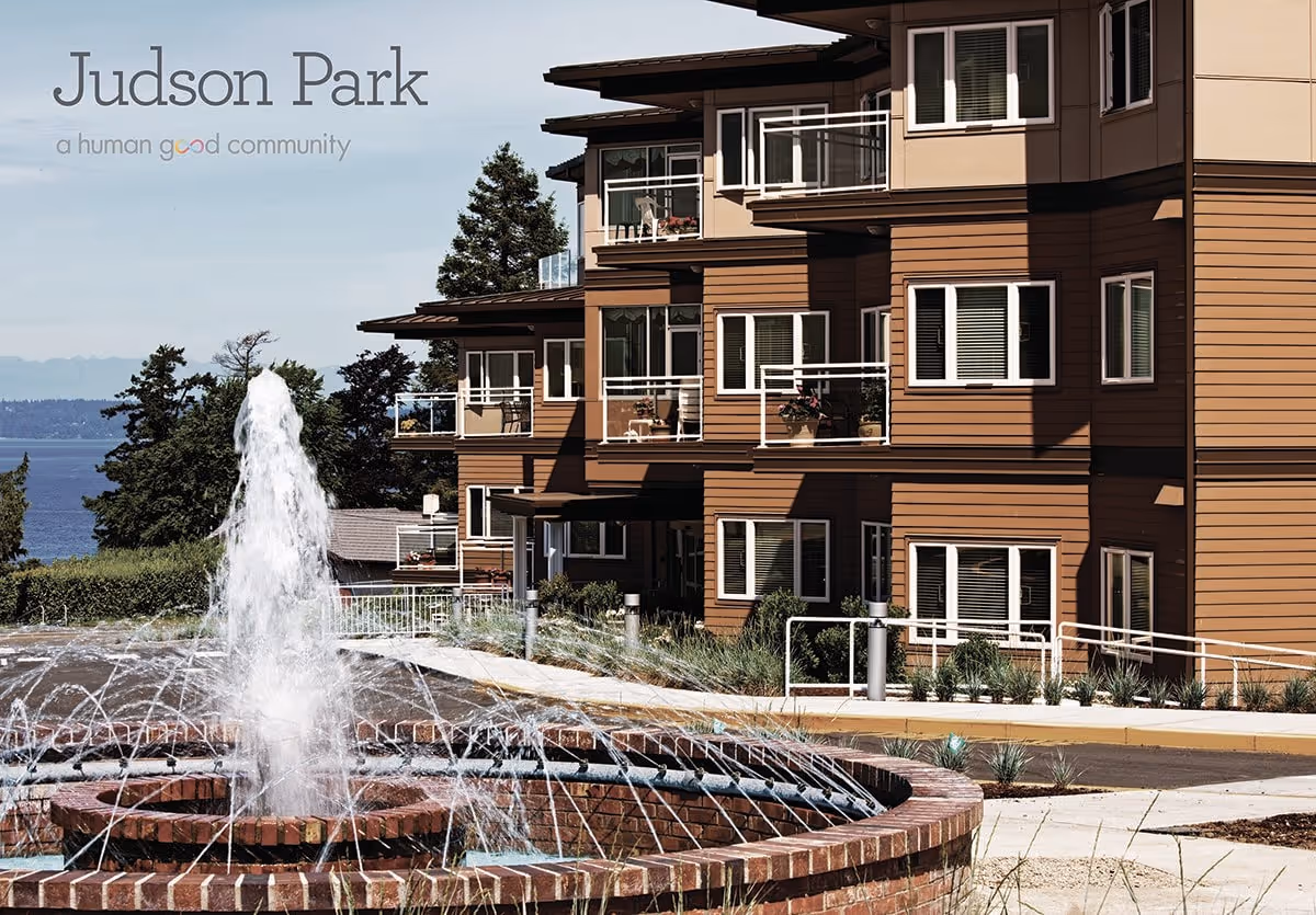 Exterior view of Judson Park Retirement Community building with multiple balconies, a large water fountain in the foreground, trees, and a body of water in the background under a clear sky.