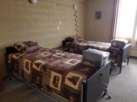 A simple bedroom with two single beds side by side, each covered with a brown patterned quilt. There is a window with vertical blinds letting in natural light, a small nightstand between the beds, and a framed picture on the wall.