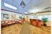 Well-lit reception lobby with a wooden front desk, rows of chairs, potted plants, framed art, and patterned carpet.