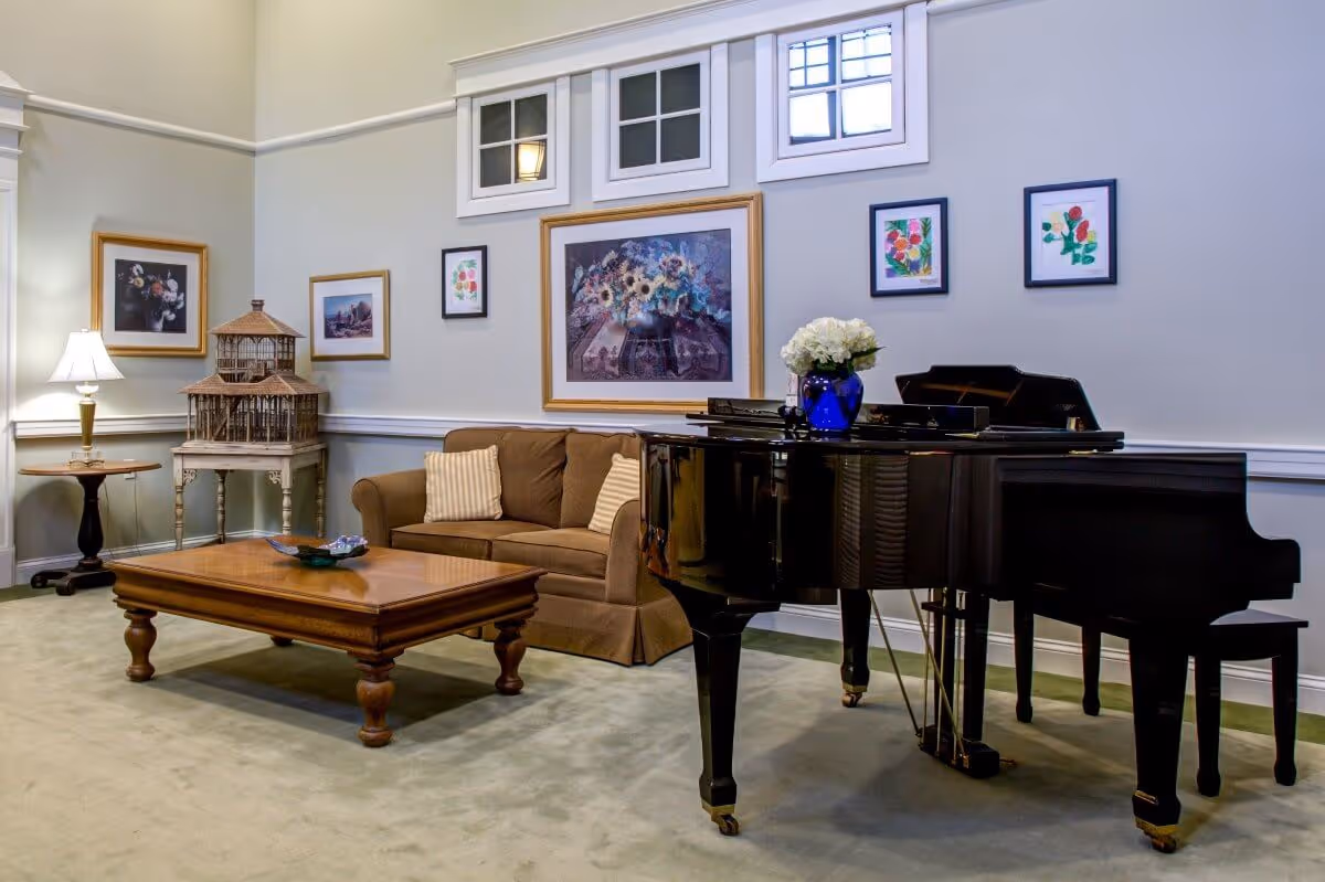 A cozy living room area with a brown sofa adorned with two striped pillows, a wooden coffee table, and a black grand piano with a blue vase holding white flowers on top. The walls are decorated with framed artwork and three small windows near the ceiling. A side table with a lamp and a decorative birdcage are also visible.