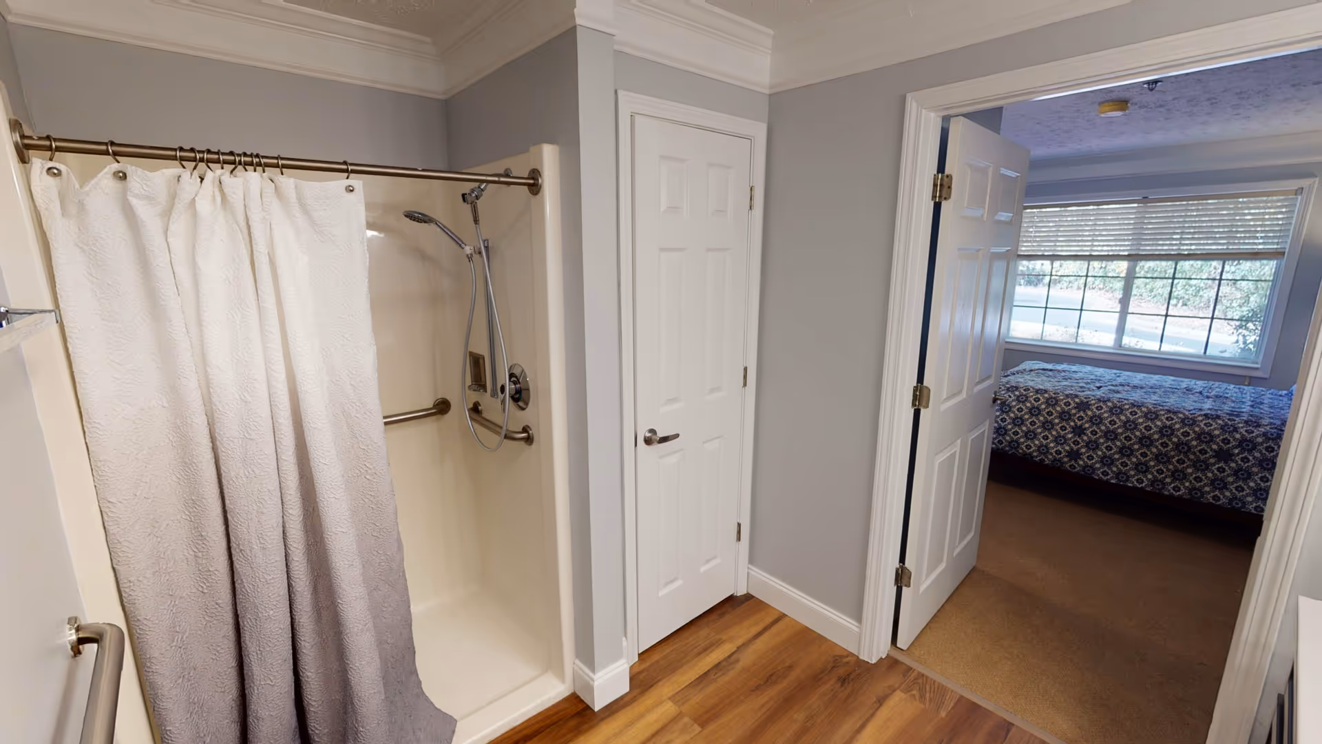 Interior view of a bathroom with a shower area featuring a white and gray gradient shower curtain, a handheld showerhead, and grab bars. The bathroom has light gray walls, white trim, and wood flooring. A white door leads to an adjacent bedroom with a bed covered in a blue patterned bedspread and a large window with blinds.