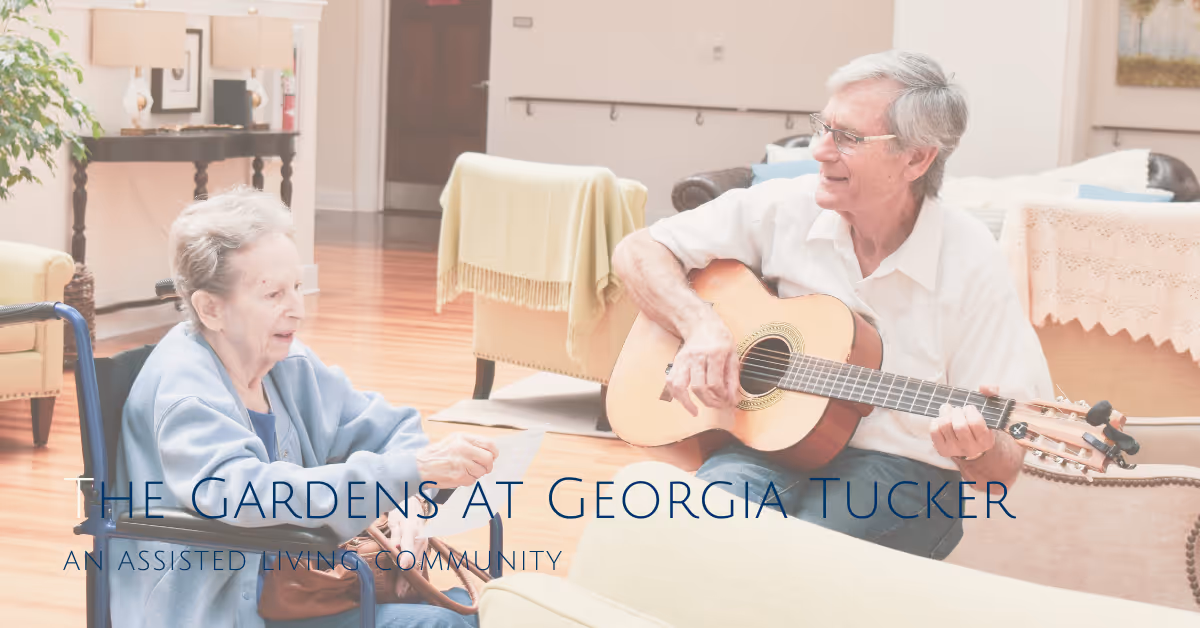 An elderly man playing an acoustic guitar while sitting on a couch, smiling at an elderly woman in a wheelchair who is holding a piece of paper. The setting is a bright, cozy living room with wooden floors, light-colored furniture, and a table with a lace cover in the background.