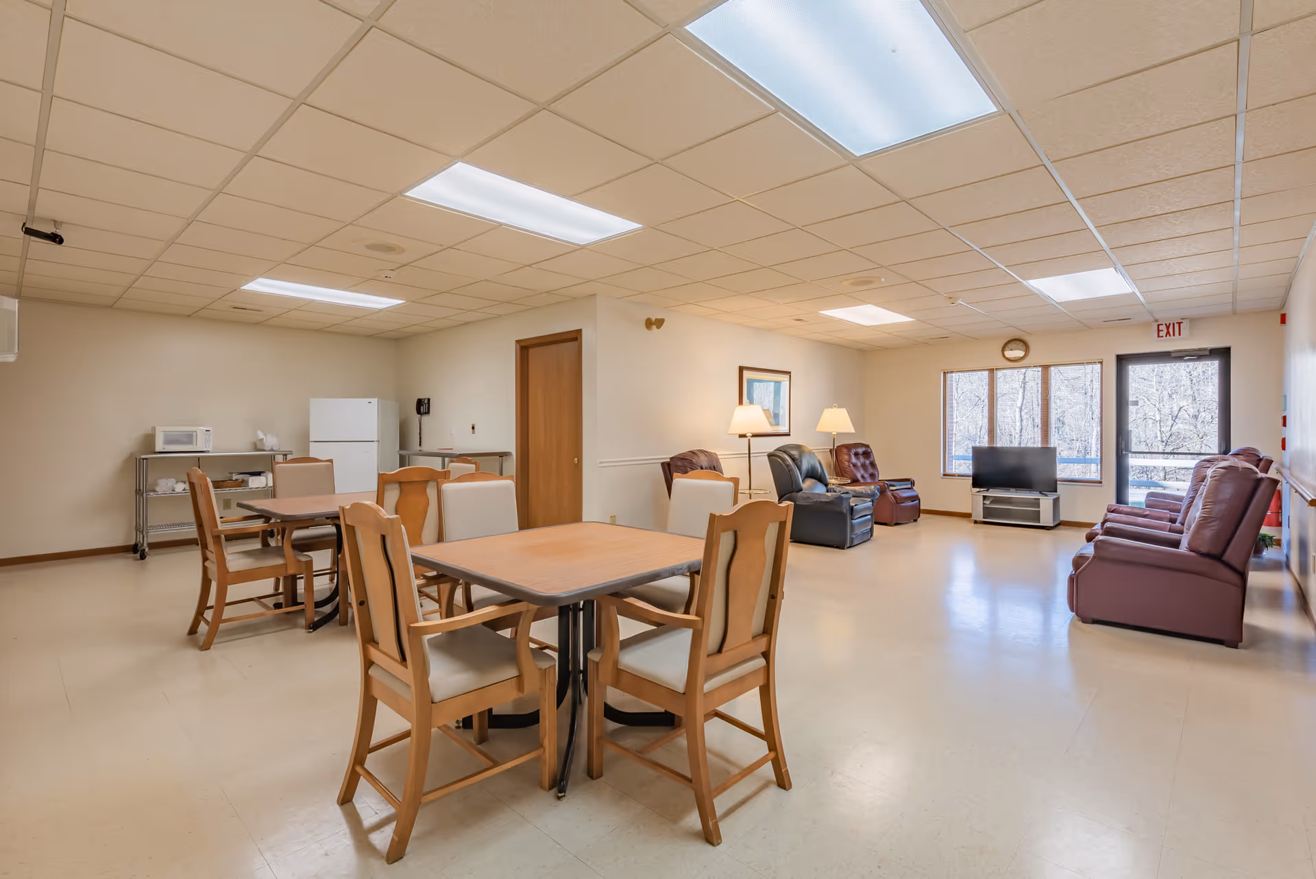 A spacious common area in a senior living facility featuring a dining section with wooden tables and chairs on the left, and a living room area with several recliner chairs, a TV on a stand, and large windows letting in natural light on the right. The room has a tiled ceiling with fluorescent lights and a door labeled EXIT near the windows.