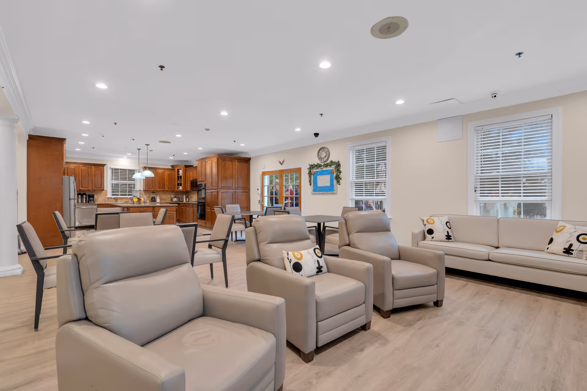 A spacious and well-lit common area in a senior living facility featuring beige leather armchairs and a matching sofa with decorative pillows. In the background, there is a kitchen area with wooden cabinets, stainless steel appliances, and a dining area with tables and chairs. The room has light-colored walls, large windows with white blinds, and recessed ceiling lights.