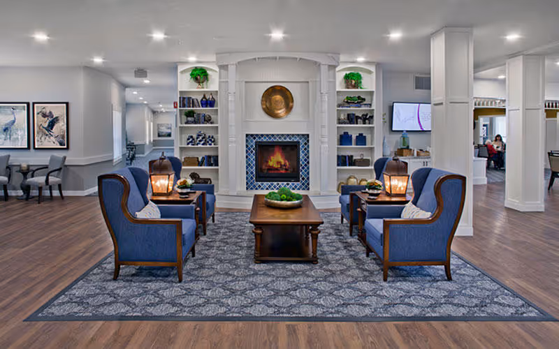 A cozy living room area with four blue upholstered armchairs arranged around a wooden coffee table on a patterned rug. Behind the seating area is a white fireplace with a decorative round brass plate above it and built-in shelves on either side holding various decorative items. The room has wood flooring and is well-lit with recessed ceiling lights. In the background, there are additional seating areas and a wall-mounted TV.