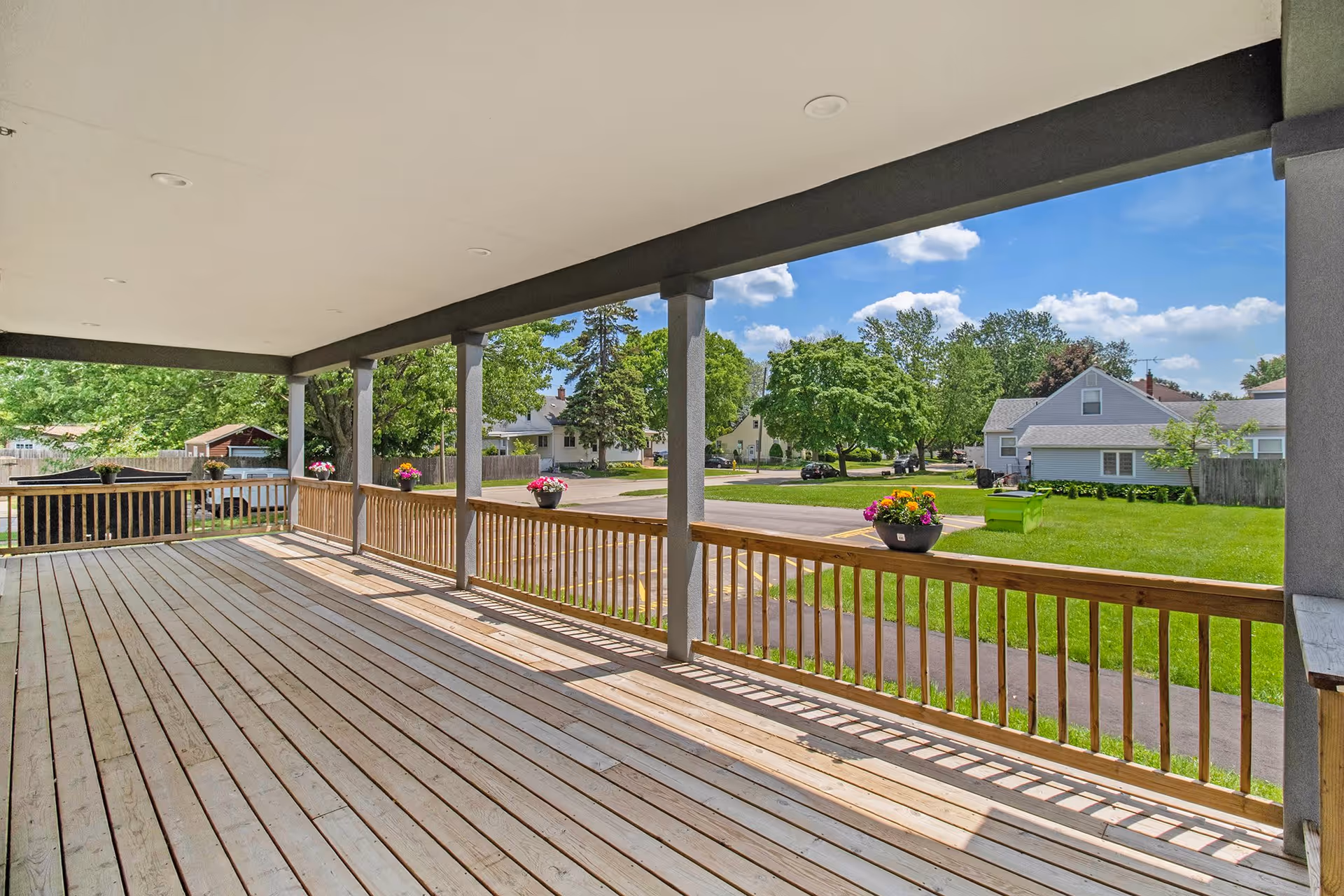 Covered wooden porch with railing and flower pots overlooking a green lawn, street, and neighboring houses under a blue sky with scattered clouds.