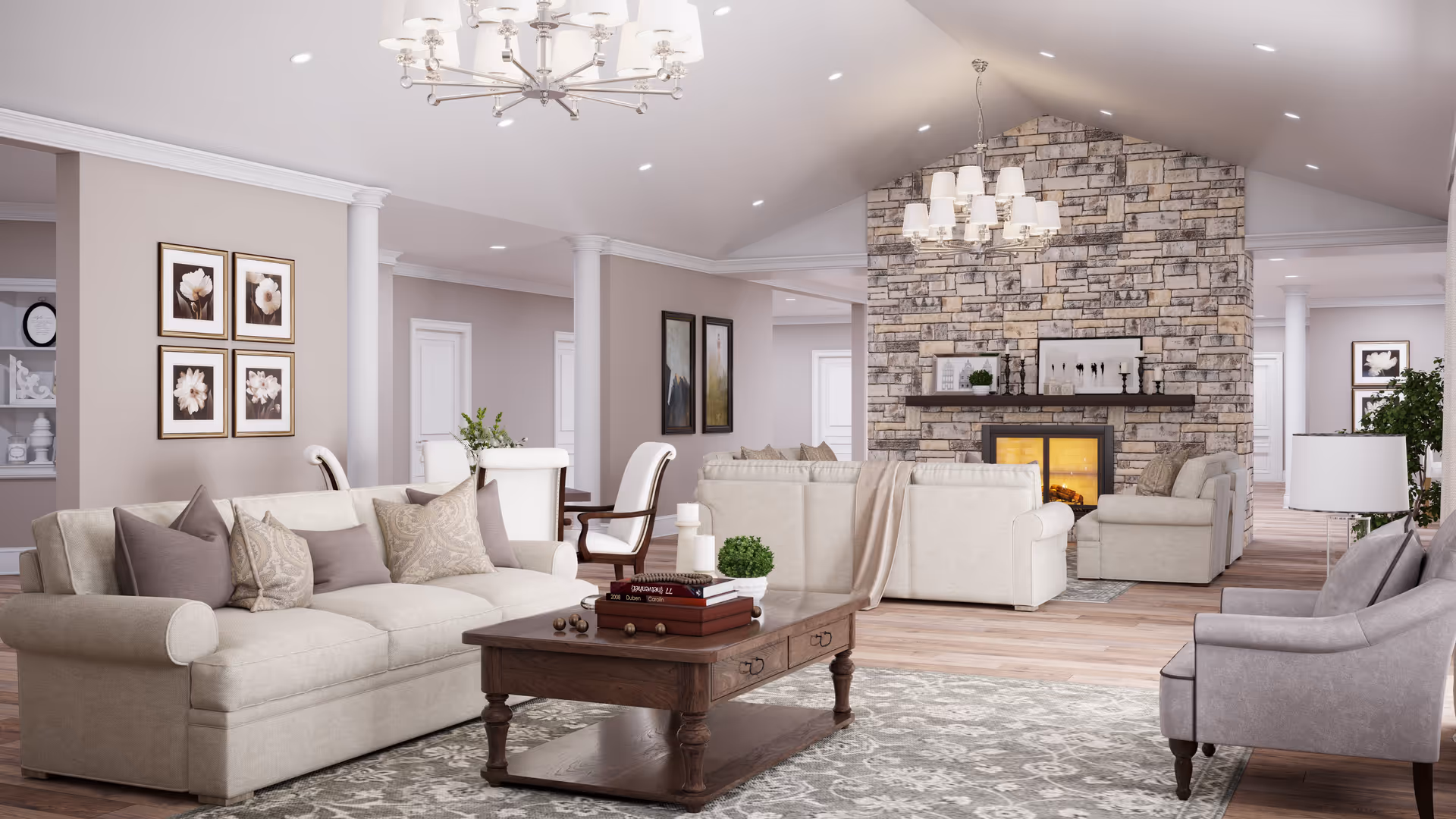 A spacious and elegant living room with beige sofas, a wooden coffee table with books and decorative items, a stone fireplace with a lit fire, and two chandeliers hanging from a vaulted ceiling. The room features light-colored walls adorned with framed artwork and a patterned area rug on wooden flooring.