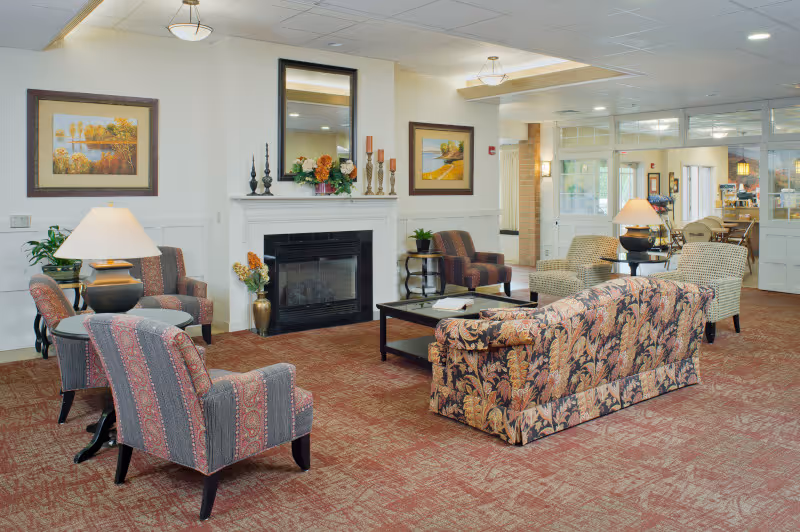 A cozy living room area in a senior living facility with patterned armchairs and a floral sofa arranged around a black coffee table and a white fireplace. The room features warm lighting from table lamps, framed landscape paintings on the walls, and a large mirror above the fireplace. The carpet is a reddish-brown color, and the space has a welcoming and comfortable atmosphere.