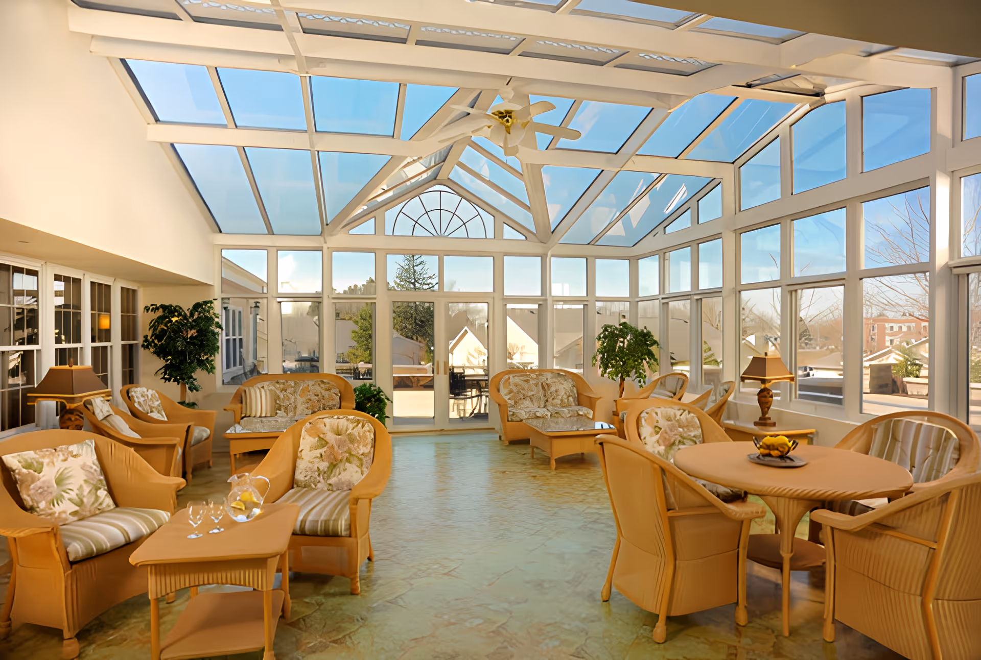 A bright sunroom with large glass windows and a glass ceiling, furnished with wicker chairs and sofas with floral cushions, small tables, lamps, and potted plants. The room has a light-colored floor and offers a view of the outdoors through the glass walls.