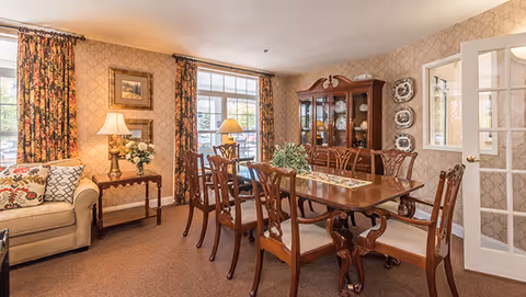 A formal dining room with a wooden table and chairs, a china cabinet, sofa, floral curtains, lamps, and patterned wallpaper.