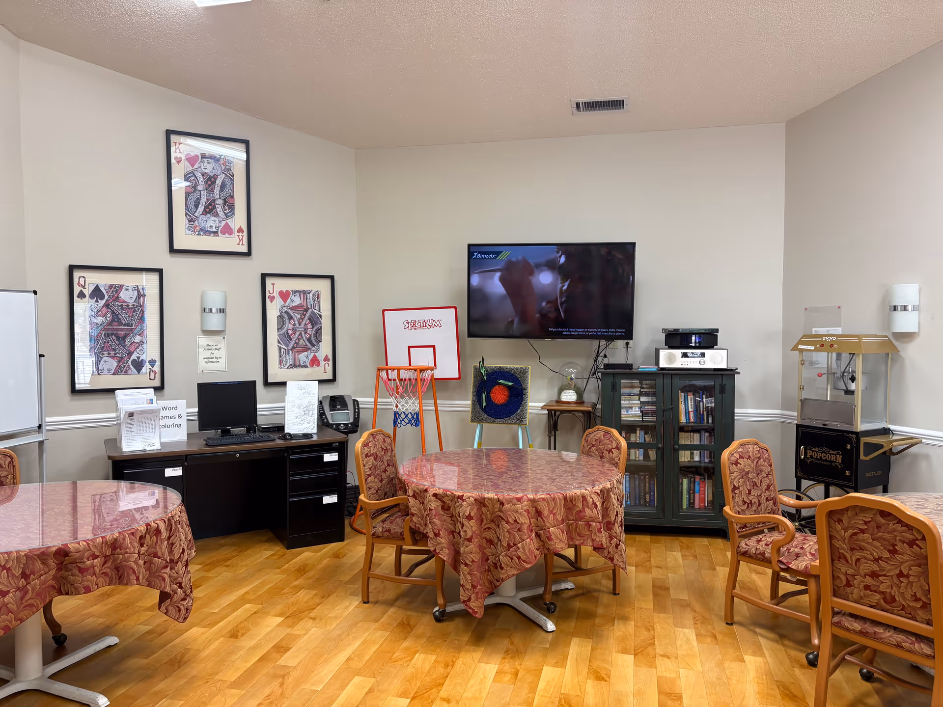 Community activity room with round tables covered in patterned tablecloths, chairs, a wall-mounted TV, bookshelves, and a popcorn machine.