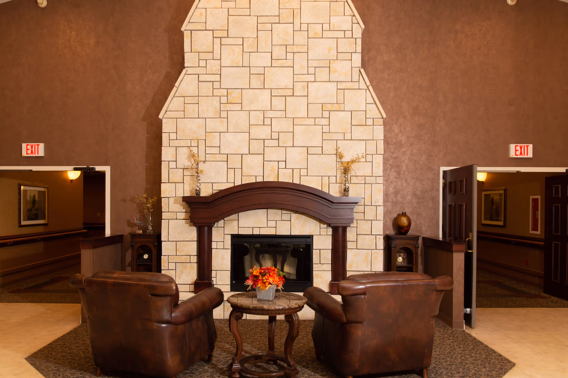 A cozy seating area with two brown leather armchairs facing a stone fireplace with a wooden mantel. A small round table with a flower arrangement is placed between the chairs. The walls are painted brown, and there are two open doorways on either side of the fireplace, each with an illuminated exit sign above.