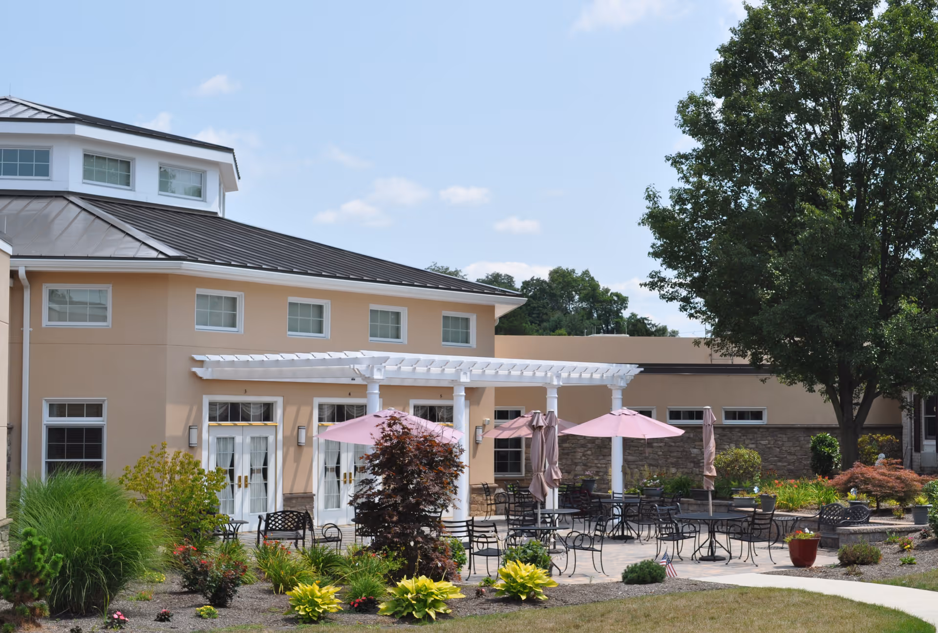 Outdoor patio area at Country Meadows Retirement Communities featuring several black metal tables and chairs with pink umbrellas, surrounded by landscaped plants and trees, adjacent to a beige building with white-framed windows and doors under a clear blue sky.