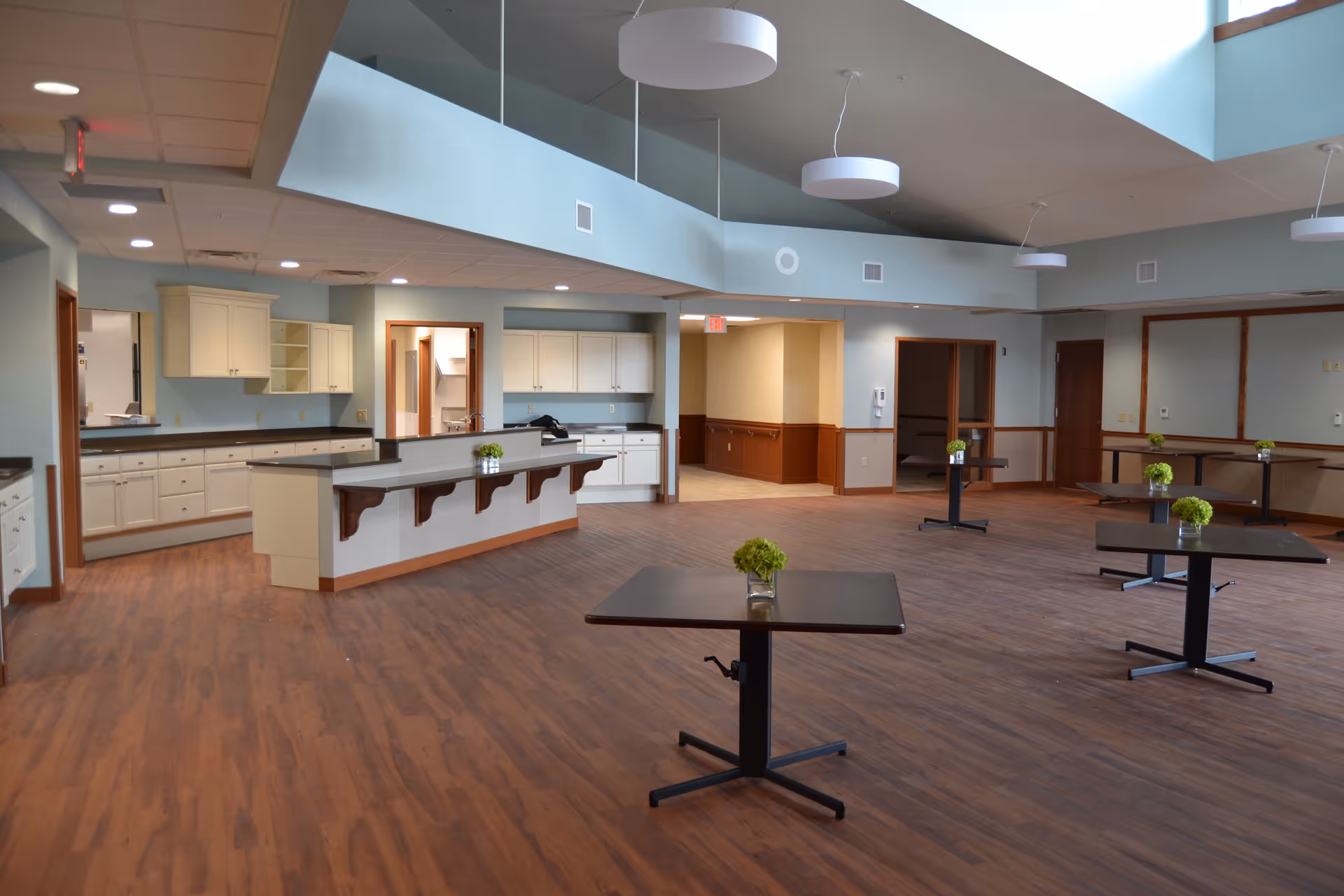 A spacious interior room with a high ceiling and modern lighting fixtures. The room features a kitchen area with white cabinets and countertops along the back wall, and several small square tables with green floral centerpieces spread across a wood-finished floor. The walls are painted light blue with wood paneling on the lower half in some areas, and there are multiple doorways leading to other rooms.