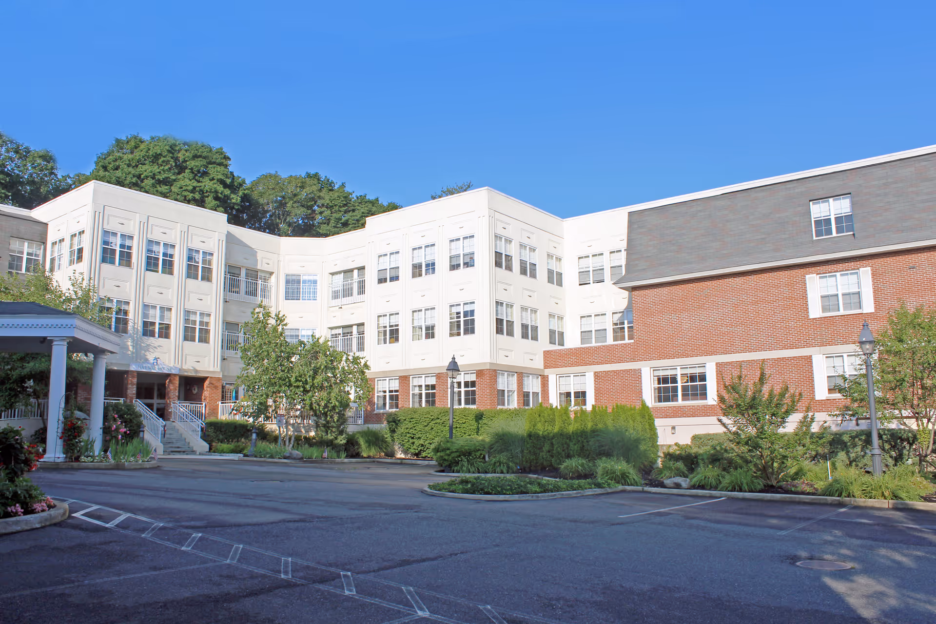 Exterior view of a multi-story assisted living facility building with white and red brick walls, numerous windows, a covered entrance, landscaped greenery, and a clear blue sky.