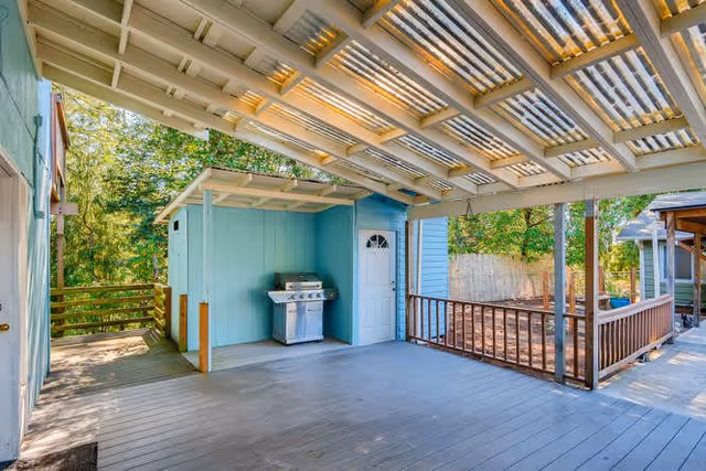Covered outdoor wooden deck with a barbecue grill, small blue storage room, and railing overlooking trees.