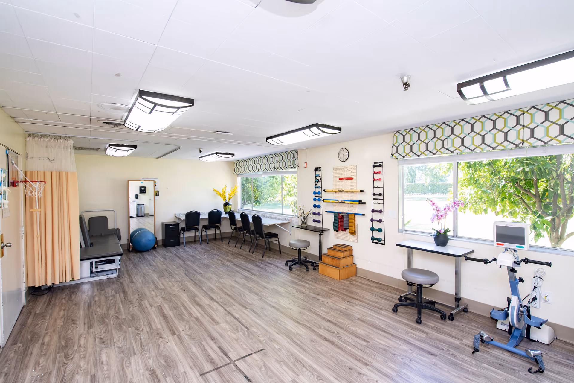 A bright and spacious rehabilitation or physical therapy room with wooden flooring, large windows with patterned valances, exercise equipment including hand weights, resistance bands, a stationary bike, and therapy steps. There are chairs lined up against the wall and a treatment table with a privacy curtain in the corner.