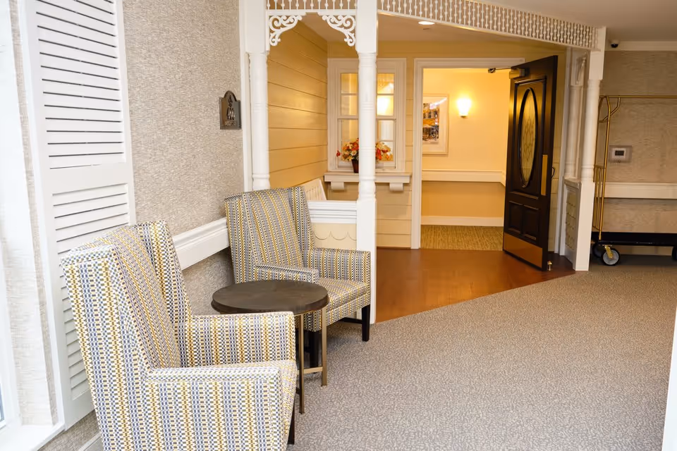 Two patterned armchairs with a small round table between them are placed in a carpeted hallway area near a doorway leading to a warmly lit room with wooden flooring. The hallway features beige walls, white trim, decorative white columns, and a coat rack with a luggage cart in the background.