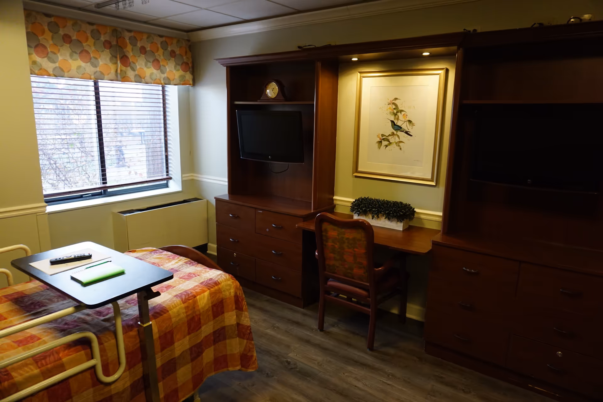 A senior living facility bedroom with a bed covered in a checkered orange and yellow blanket, a small overbed table with a remote and notepad, a window with blinds and a patterned valance, two wooden dressers with mounted flat-screen TVs, a wooden desk with a chair, a framed bird artwork on the wall, and a small potted plant on the desk.