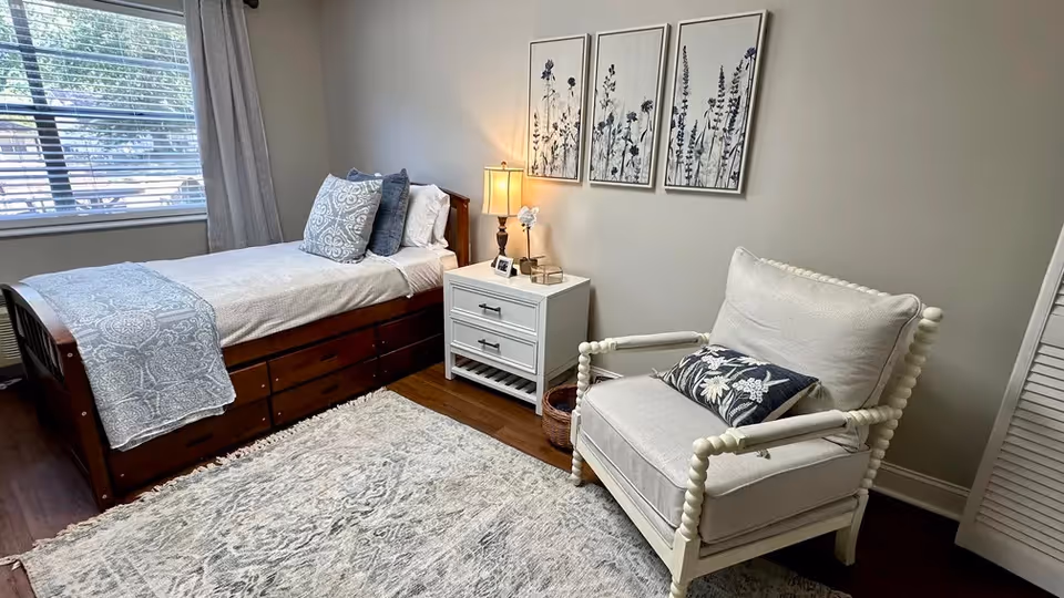 A cozy bedroom with a single wooden bed dressed in white and blue bedding, a white nightstand with a lamp and decorative items, a white cushioned armchair with a floral pillow, three framed botanical prints on the wall, a large window with blinds and curtains, and a patterned area rug on a wooden floor.