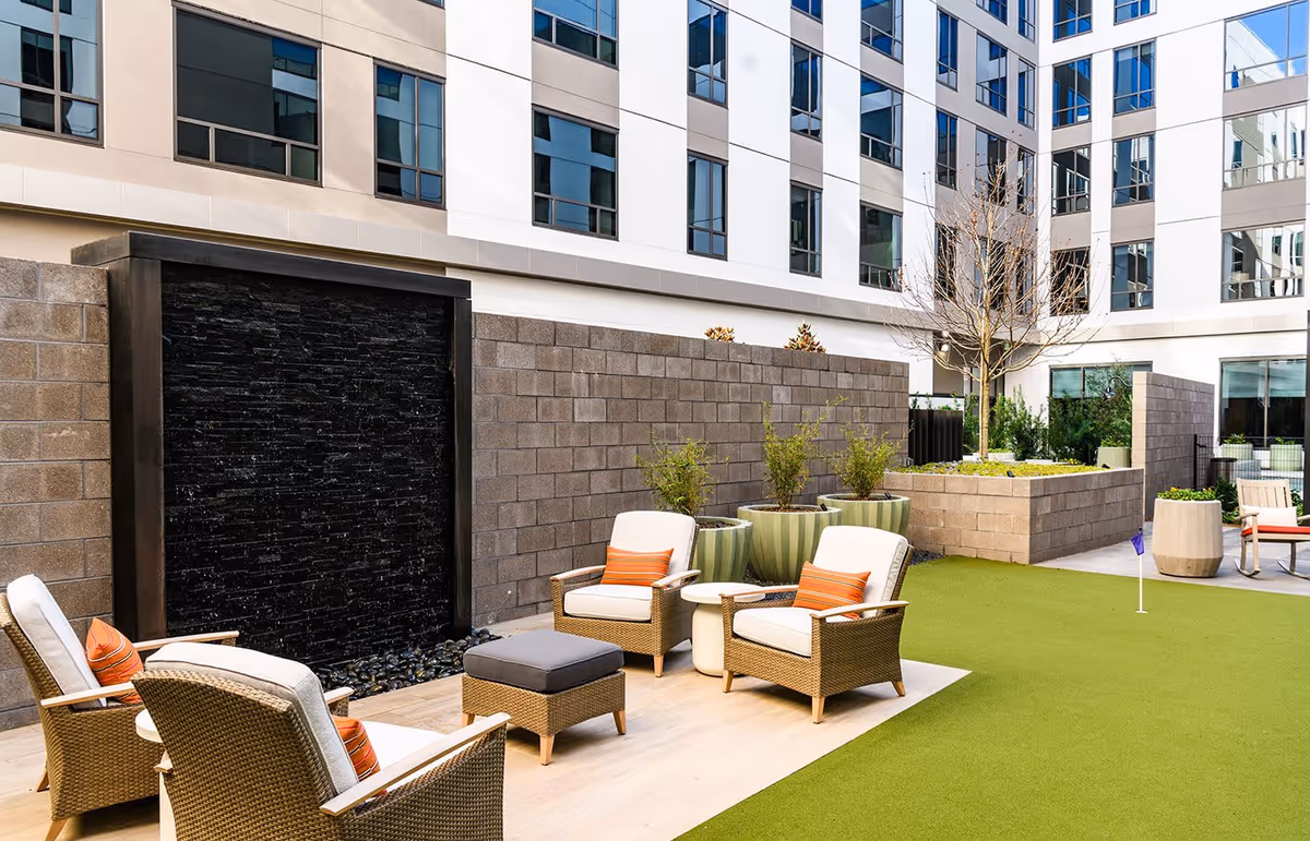 Outdoor courtyard area at Clearwater at The Heights featuring wicker chairs with cushions and orange striped pillows, a small white table, a black water feature wall, potted plants, a putting green with a small flag, and a multi-story building with many windows in the background.