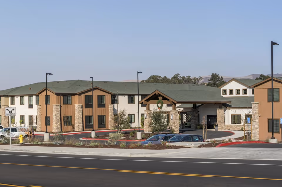 Front exterior of a two-story senior living building with a covered entrance, parking area, and landscaped driveway.