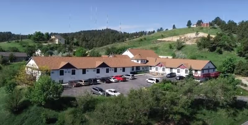 Aerial view of a single-story assisted living facility with a red-tiled roof surrounded by greenery and hills. Several cars are parked in the parking lot in front of the building under a clear blue sky.