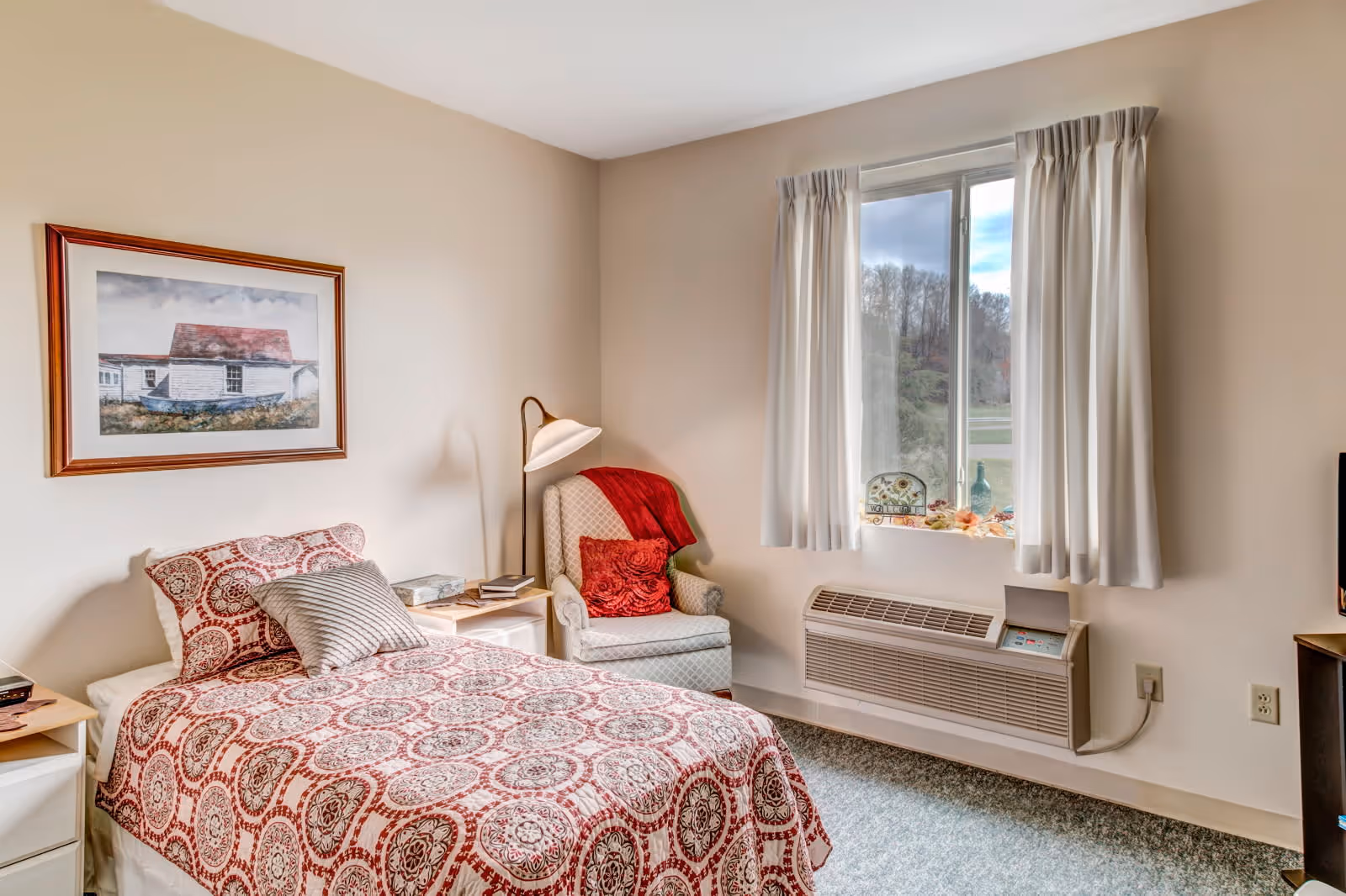 A cozy bedroom with a single bed covered in a red and white patterned bedspread, a white armchair with a red pillow and throw, a floor lamp, a window with white curtains, and a framed picture on the wall.