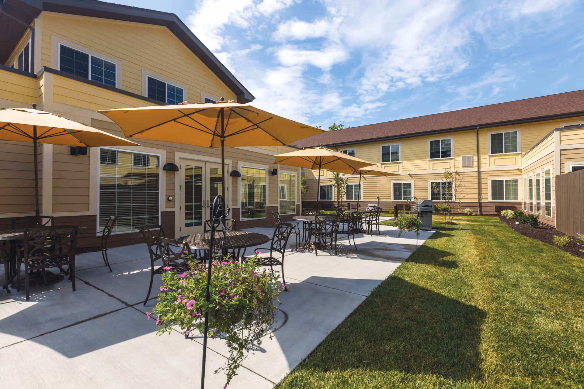 Outdoor patio area at Dougherty Ferry Assisted Living and Memory Care with several round metal tables and chairs under large yellow umbrellas, adjacent to a two-story yellow building with multiple windows. The patio is surrounded by a well-maintained lawn and some small plants and flowers.