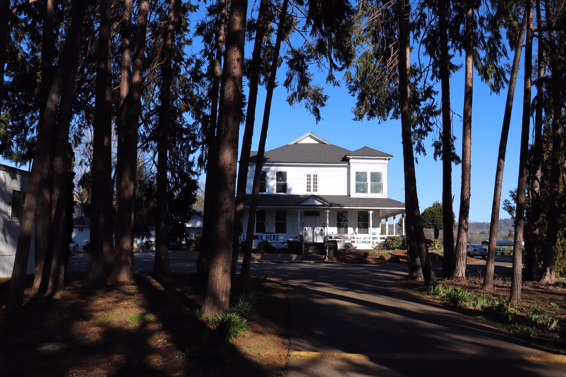 A white two-story building with a wraparound porch, partially obscured by tall trees in the foreground. The building is set against a clear blue sky and is surrounded by a driveway and some landscaping.