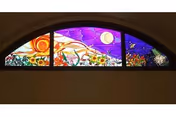 A colorful stained glass window with a semi-circular arch shape, featuring a vibrant design of sunflowers, a bright sun, a crescent moon, stars, and various abstract patterns in shades of purple, orange, yellow, and green.