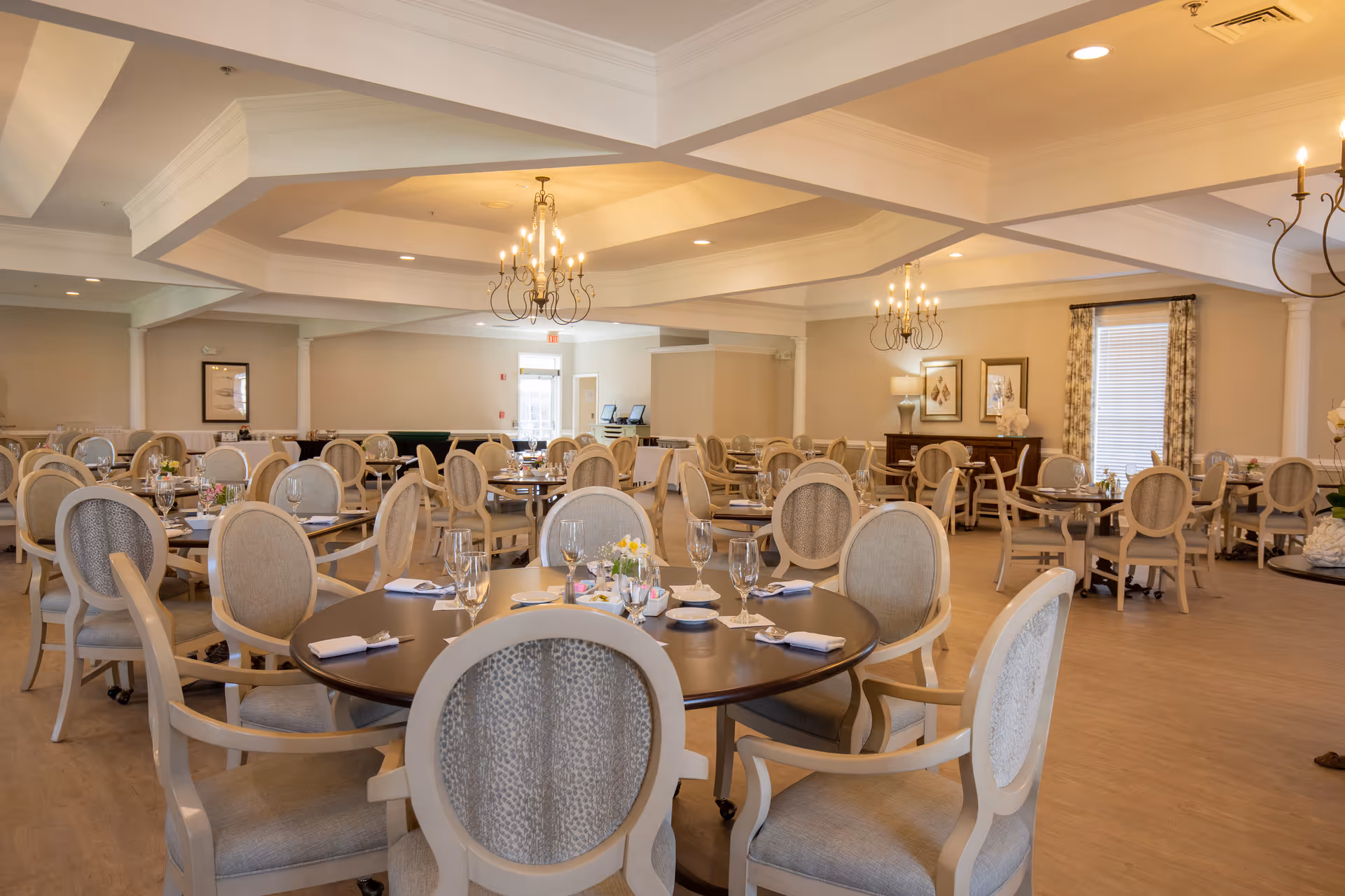 Spacious, elegantly furnished dining room with round tables set for service and chandeliers overhead.