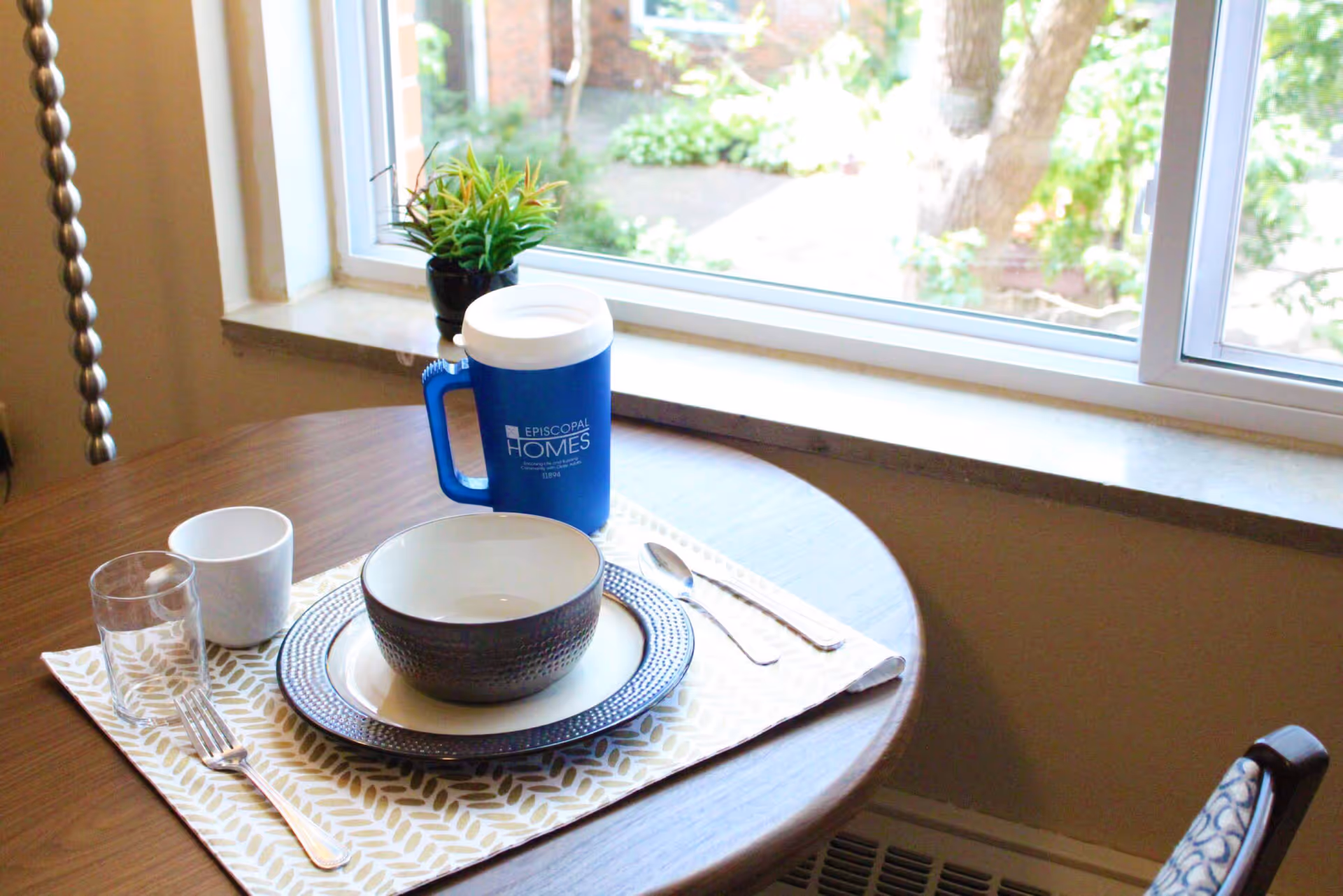 A small round wooden dining table set for one with a bowl, plate, fork, spoon, glass, and cup on a patterned placemat. A blue insulated cup with the text 'Episcopal Homes' is also on the table. The table is positioned next to a window with a view of greenery outside, and a small potted plant is on the windowsill.
