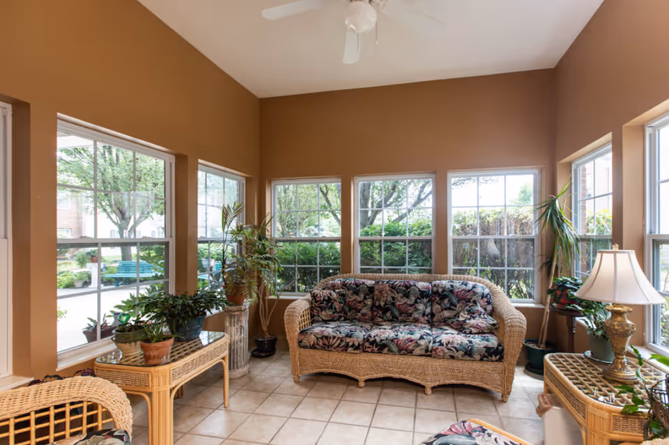 A sunlit sitting area with large windows on three sides showing green trees and bushes outside. The room has tan walls and tiled floor. There is a wicker sofa with floral cushions, a wicker chair, a wicker coffee table with several potted plants, and a wicker side table with a decorative lamp and more plants. A white ceiling fan is mounted above.