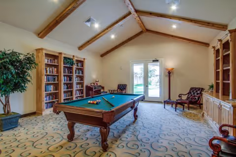 A spacious recreational room with a pool table in the center, surrounded by wooden bookshelves filled with books. The room has a high ceiling with exposed wooden beams, a patterned carpet, a potted plant, and a door leading outside. There are also a few chairs and a floor lamp near the door.