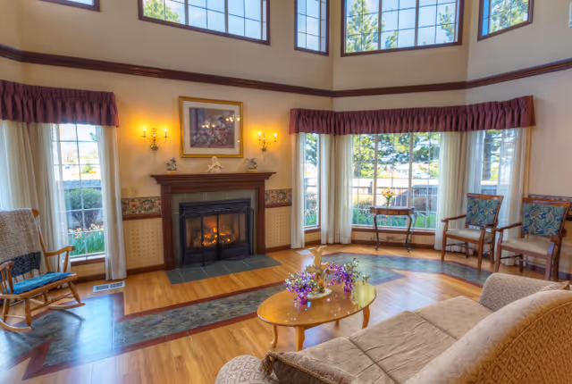 Bright, two-story common living room with tall windows, a lit fireplace, sofa and chairs around a coffee table with flowers.