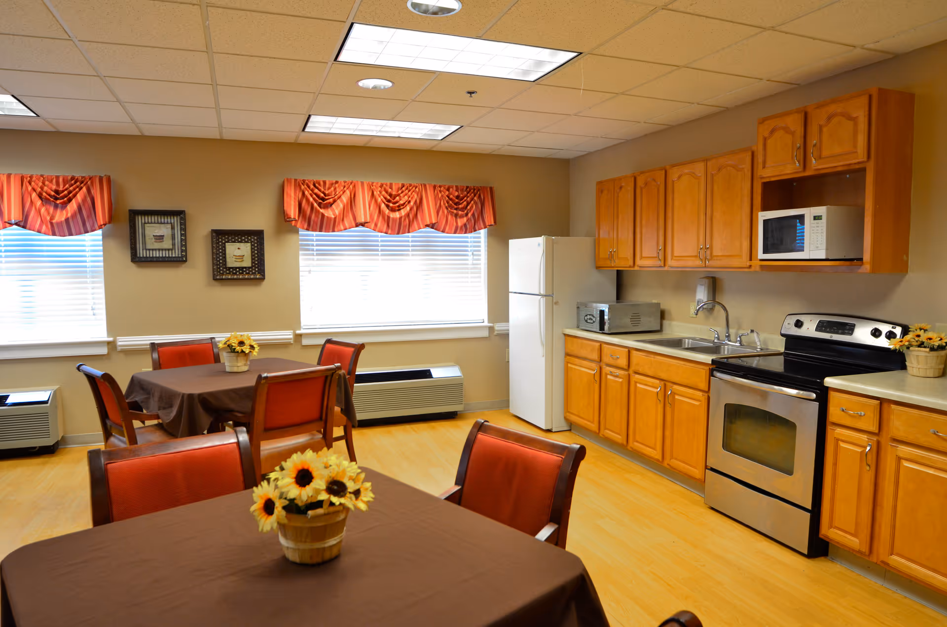 A bright kitchen and dining area with wooden cabinets, a white refrigerator, stainless steel oven, microwave, and toaster. There are two tables covered with brown tablecloths, each with a small flower arrangement featuring sunflowers. The room has large windows with red valance curtains and framed pictures on the wall.