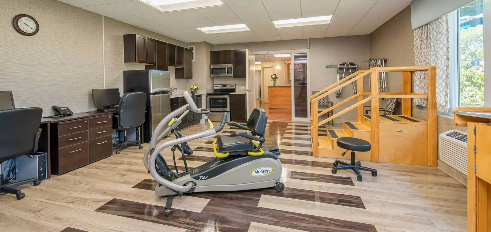 A multi-purpose room in a senior living facility featuring exercise equipment, two office desks with chairs and computers, a kitchenette with stainless steel appliances, and a wooden staircase with handrails. The room has wood-patterned flooring and a large window with patterned curtains allowing natural light to enter.