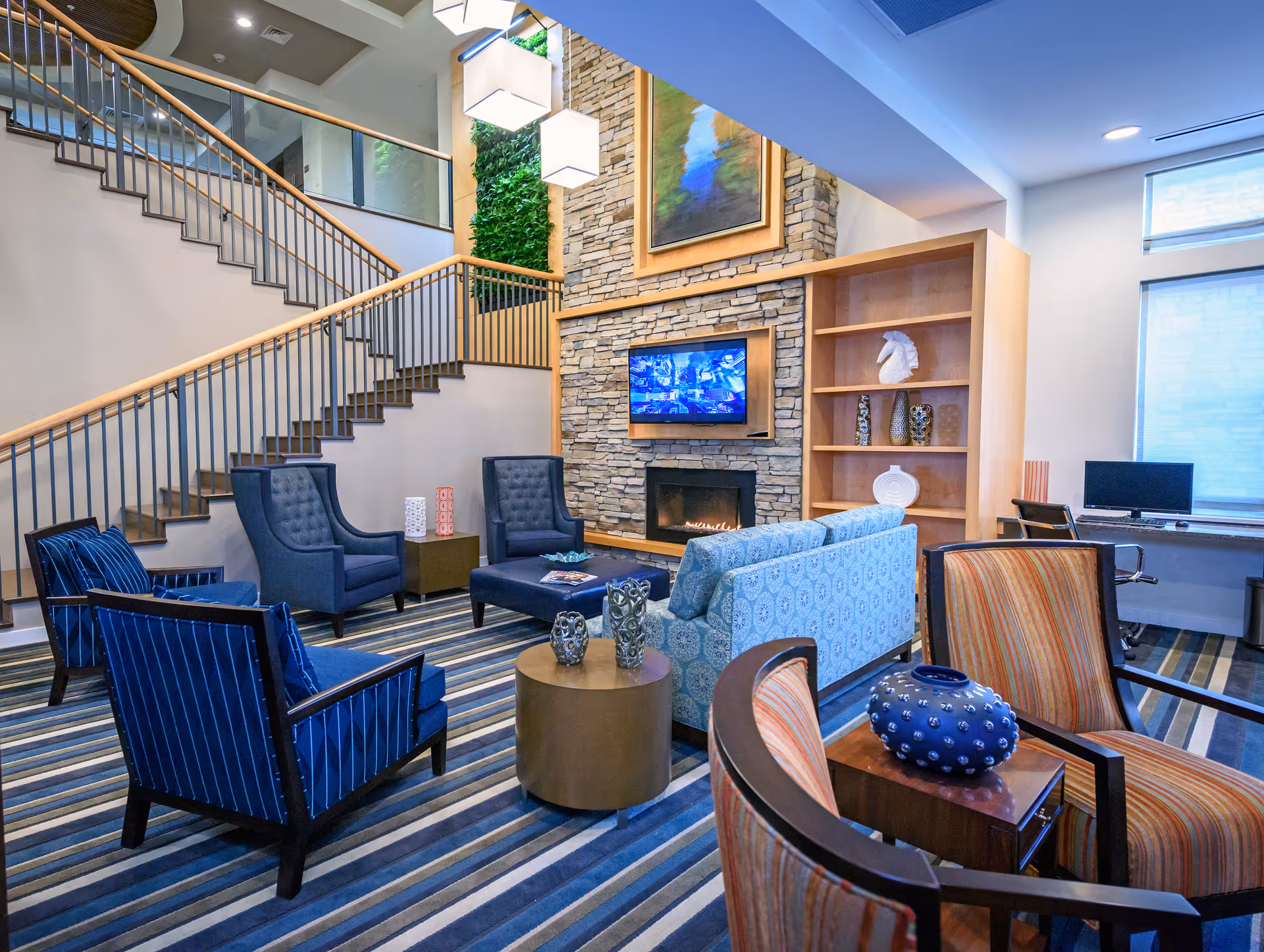 A modern living room area with a striped carpet, multiple armchairs in blue and orange patterns, a blue patterned sofa, a round coffee table with decorative items, a stone fireplace with a mounted TV above it, and a wooden bookshelf with decorative pieces. A staircase with metal railings and wooden handrails is visible on the left side, and a desk with a computer is seen near a window on the right.