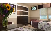 Furnished senior living bedroom with two beds, dark wardrobe, flower bouquet, and wall-mounted TV.