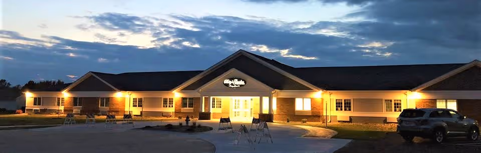 Exterior view of a single-story senior living facility building named Gardens at Kewanee during dusk, with lights illuminating the entrance and windows. A car is parked on the right side near the building.