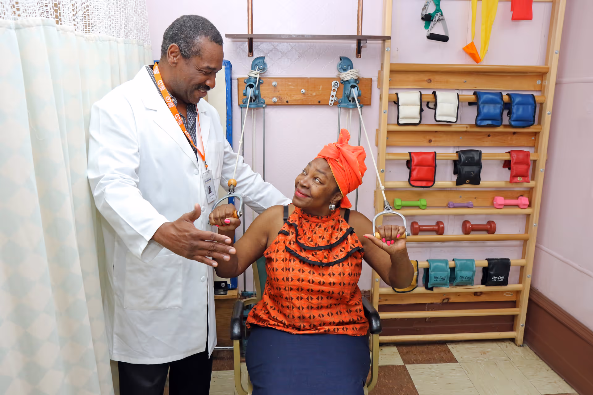 A healthcare professional in a white coat assists a seated woman in an orange patterned top and headwrap with arm exercises using pulley equipment in a therapy room with various weights and exercise bands on the wall.