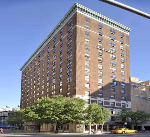 Large multi-story brick residence building on a tree-lined city corner under a clear blue sky.