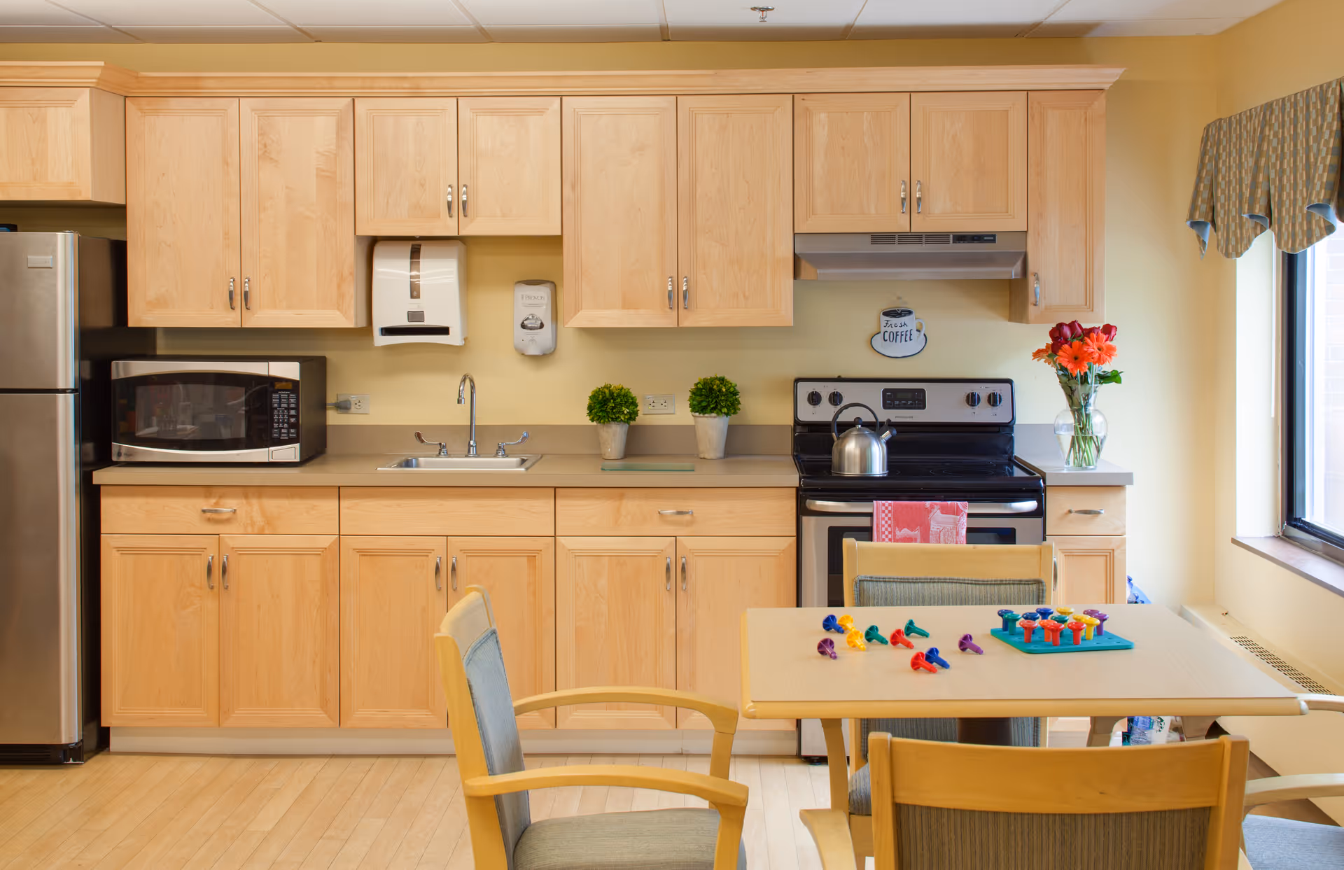 Bright senior living facility kitchen with light wood cabinets, stainless steel appliances, and a dining table with chairs.