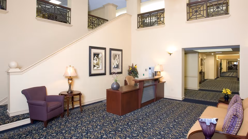 A bright and spacious senior living community lobby area with a patterned blue carpet, a wooden reception desk with a chair, a purple armchair next to a small round table with a lamp, and a beige sofa with purple pillows. The walls are light-colored with framed floral artwork, and there is a staircase with a railing leading to an upper floor. The hallway extends further into the building.