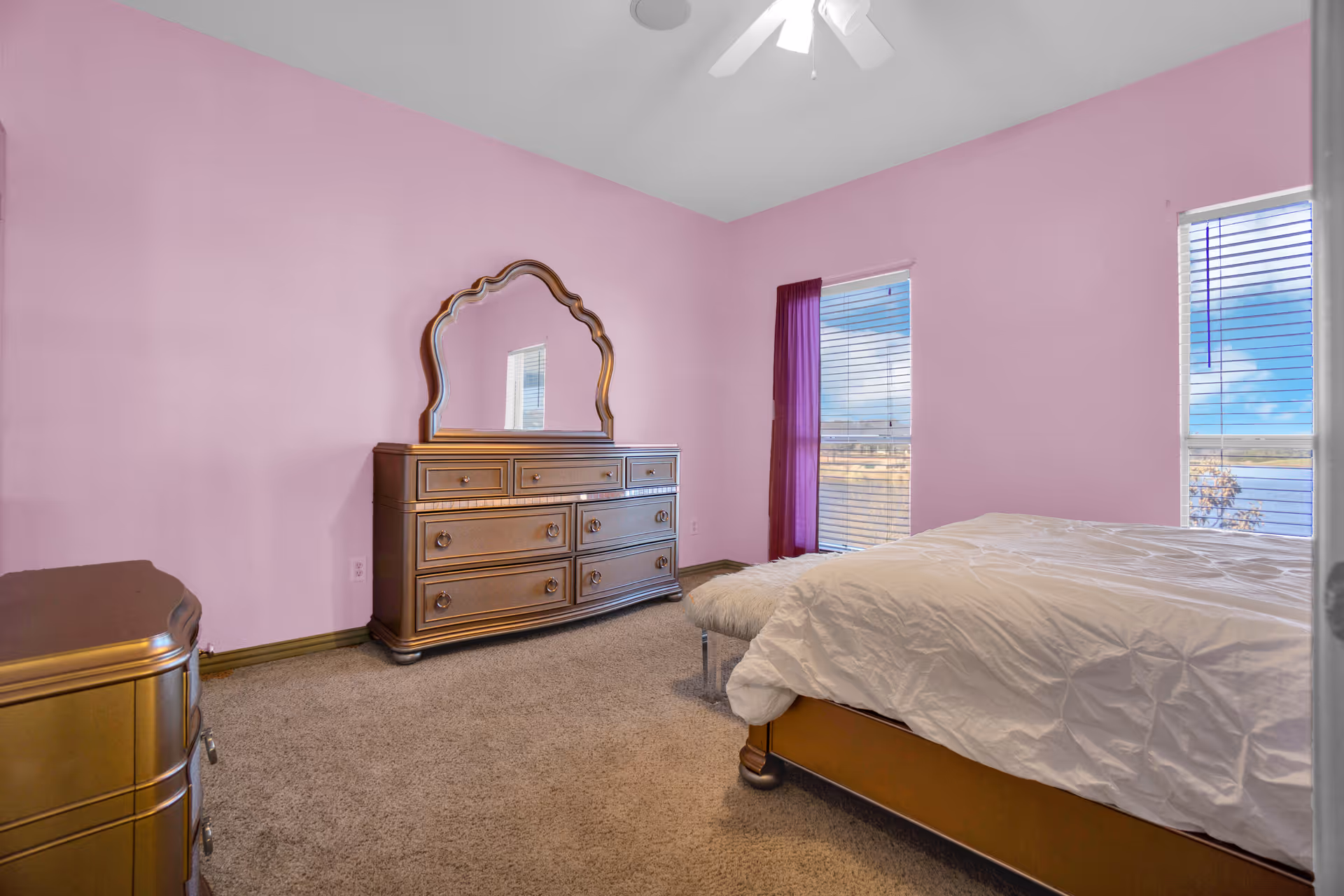 A bedroom with pink walls, beige carpet, and two windows with blinds and a purple curtain. The room contains a wooden dresser with a large decorative mirror, a matching nightstand, and a bed with a white comforter and a furry bench at the foot.