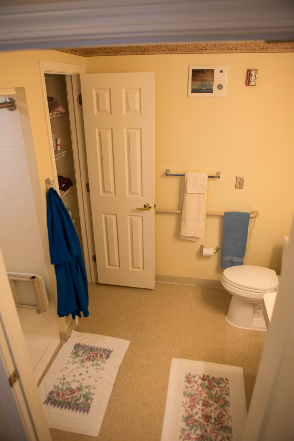 A small bathroom with a toilet, towel racks, a shower, and floral bath mats on the floor.